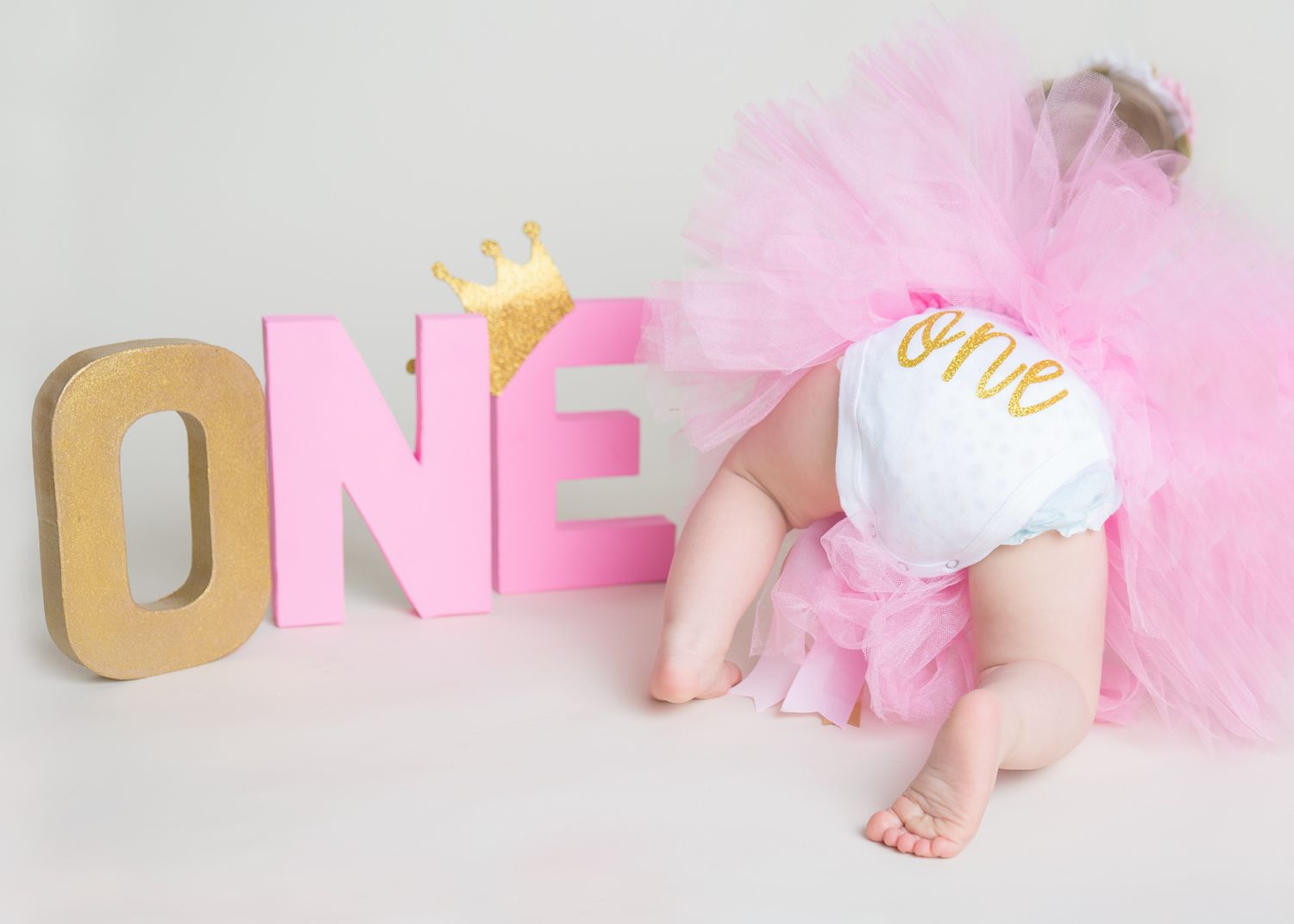 Baby in pink tutu dress crawls across white floor with gold number one decorations for first birthday photo shoot - milestone cake smash