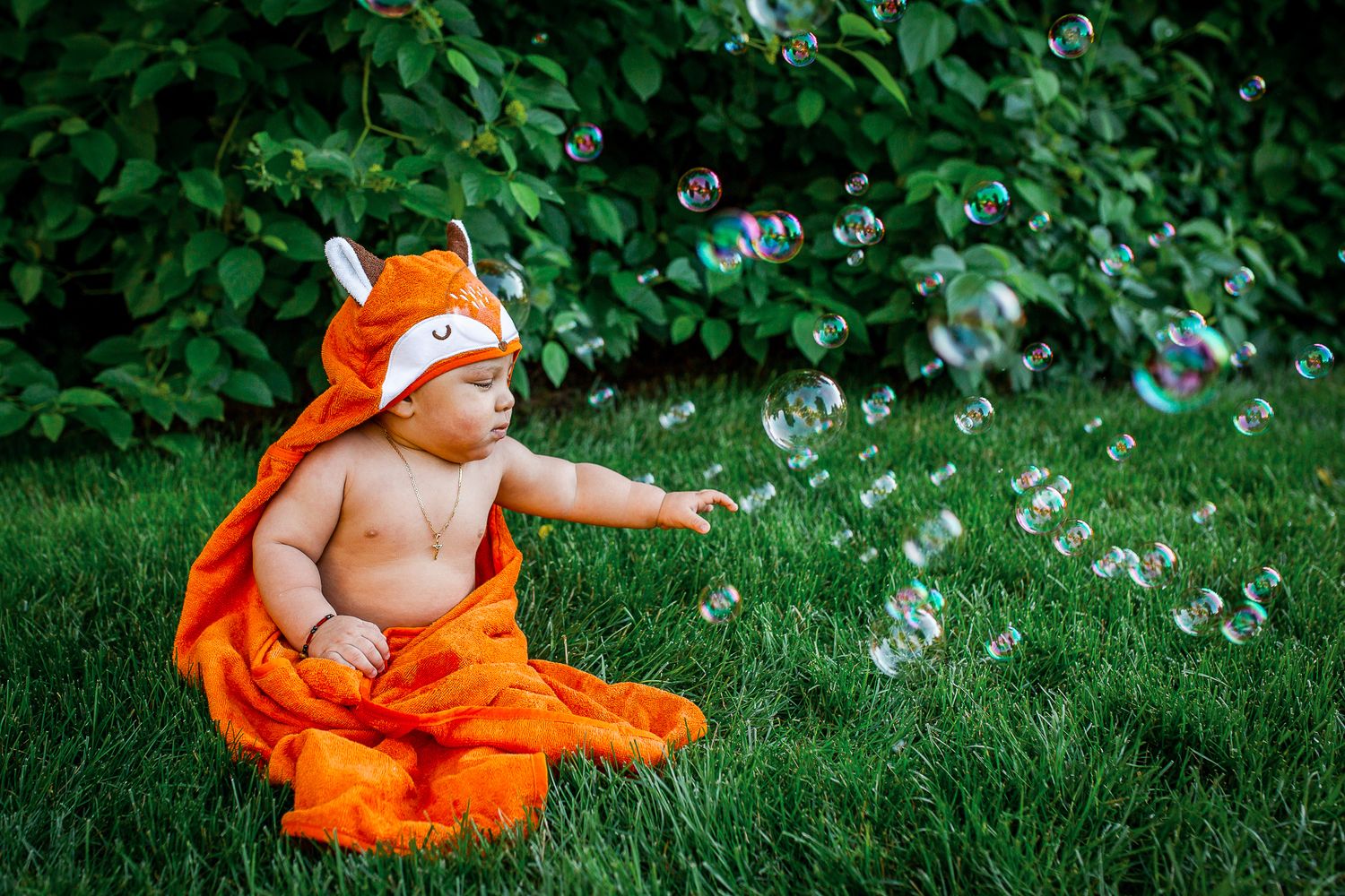 Baby wearing orange fox towel sits on grass reaching for floating soap bubbles in a garden setting - 9 month milestone photos Boise Idaho