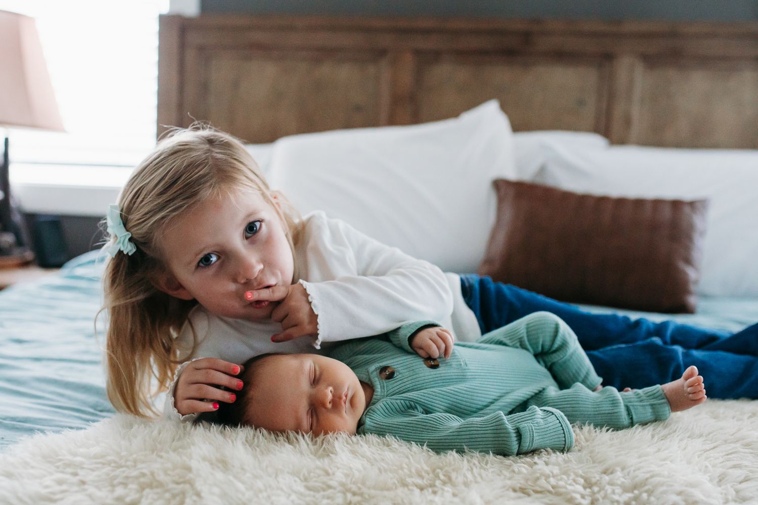 A young toddler lying on a bed behind a sleeping newborn, holding a finger to her lips as if shushing to keep the baby quiet - Boise photos