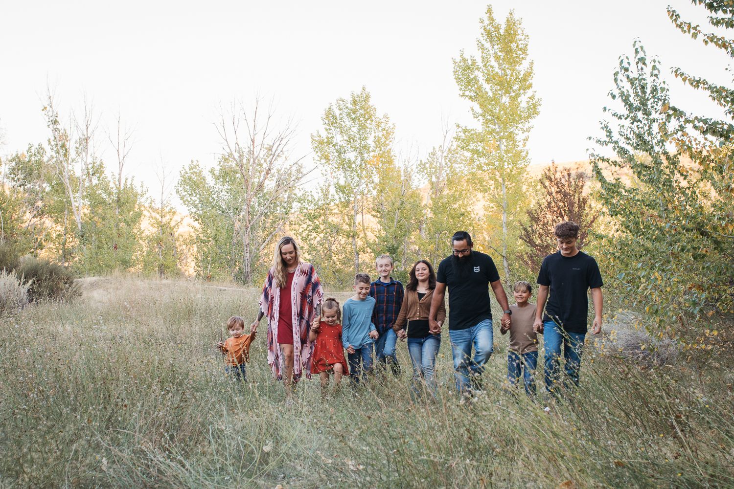 A large family walks together through a golden field during sunset while trees sway in the background - lifestyle family photography Boise