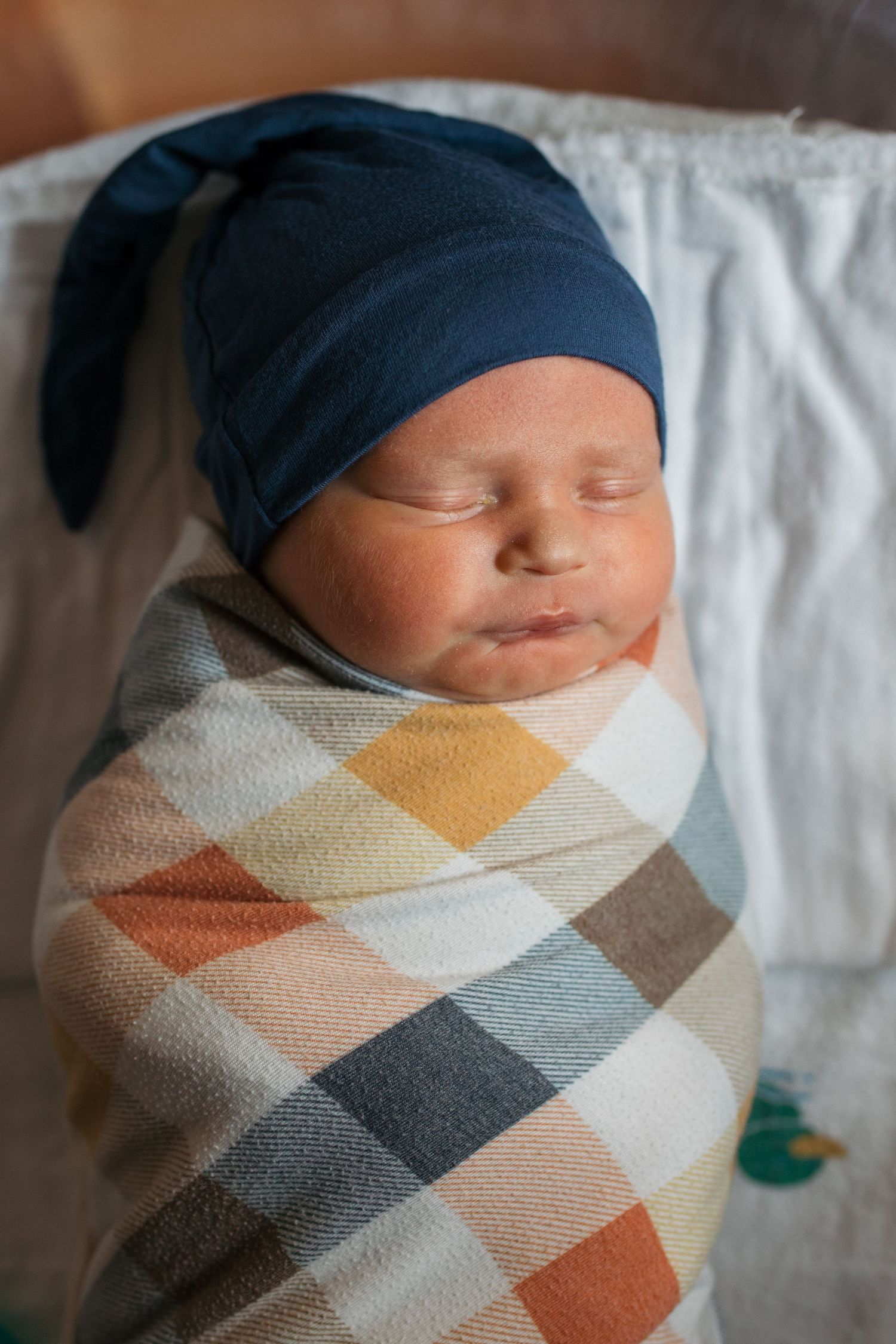 A sleeping baby wrapped in a geometric patterned blanket wearing a blue hat on white bedding - Fresh 48 session in Boise, Idaho
