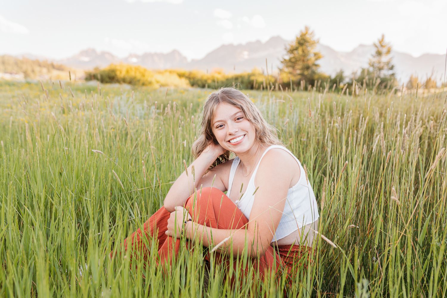 A girl sitting in tall green grass during a golden sunset with mountains visible in the background - Senior photos Caldwell Idaho