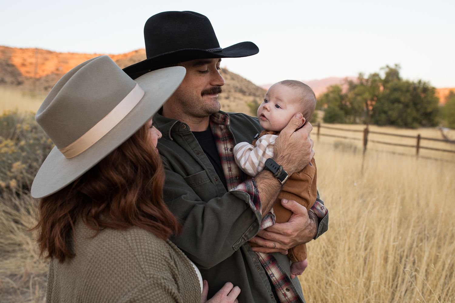 A couple wearing cowboy hats share a tender moment with their baby in a golden field at sunset - lifestyle family photos in Boise
