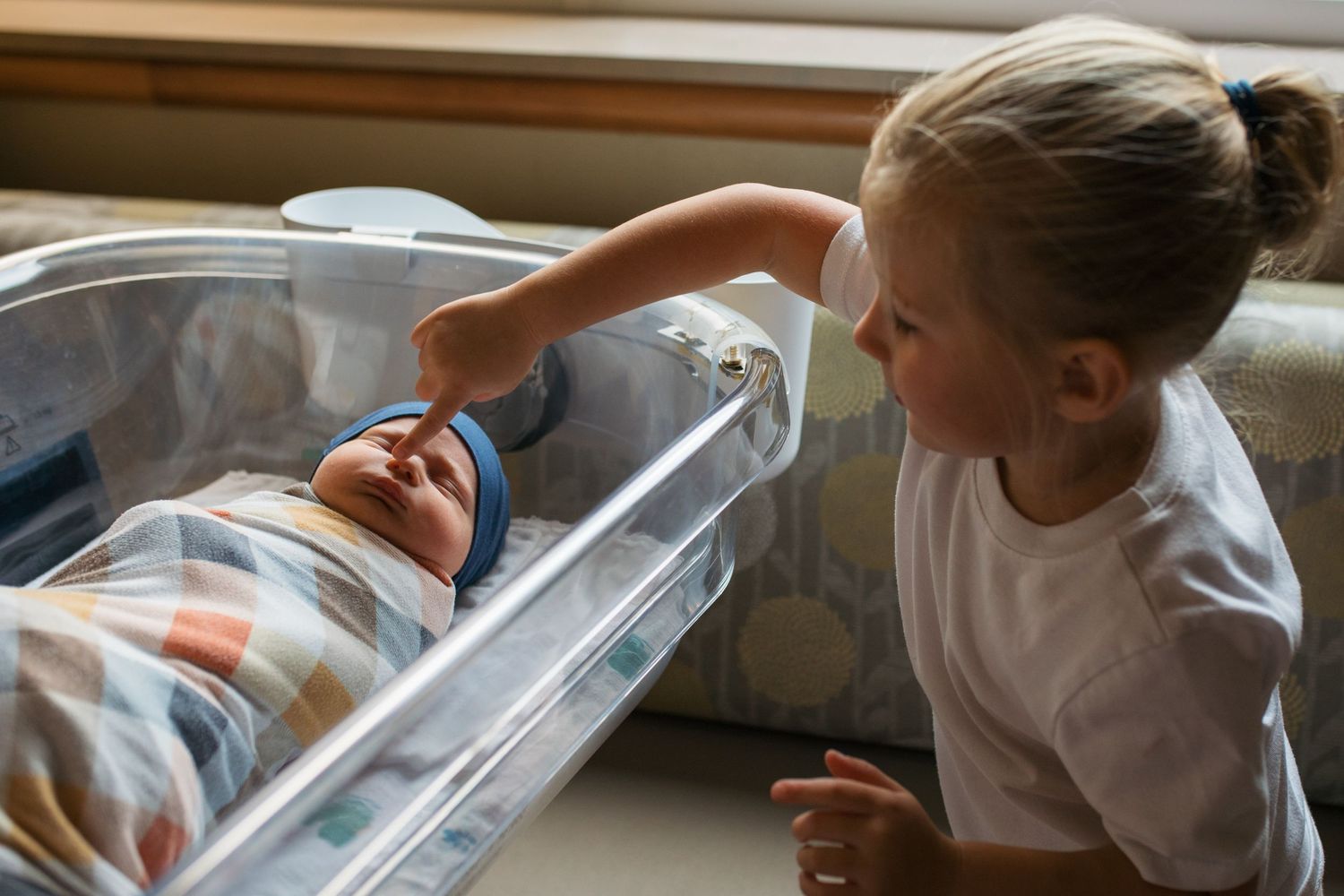 Newborn baby swaddled, sleeping in a hospital bassinet next to a window, while his older toddler sibling gently reaches in to touch him