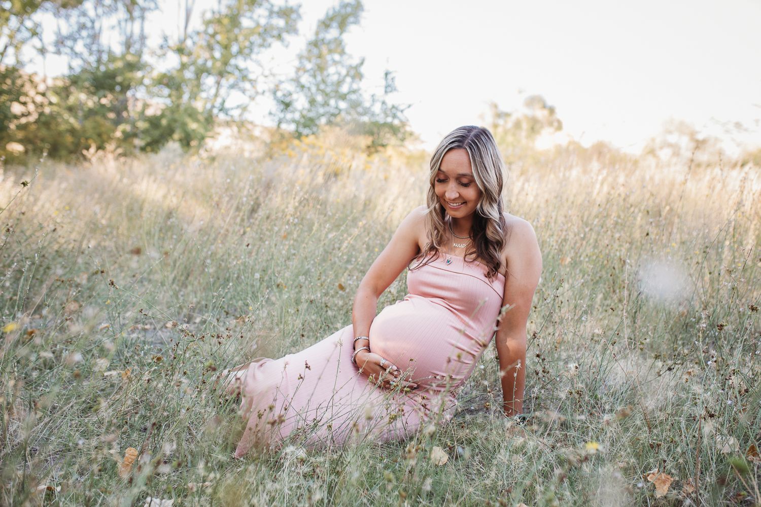 Pregnant woman in a flowing pink dress standing in the foothills of Boise, gently caressing her baby bump. Maternity session in Boise, Idaho