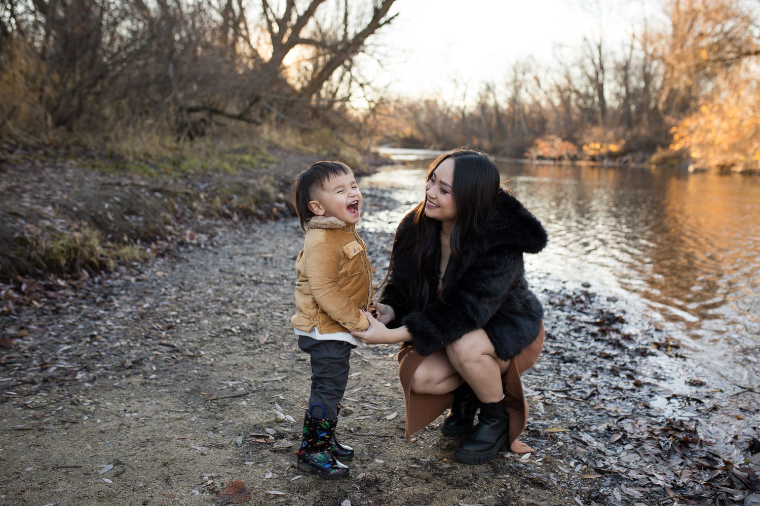 Mom smiling at her laughing toddler by the river at Reid Merrill Park in Eagle, Idaho during warm winter sunset