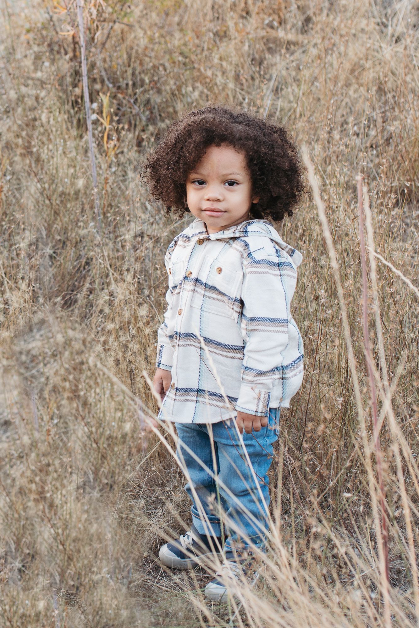 Small child wearing plaid flannel shirt and jeans stands among tall dried grass in autumn field - lifestyle family photos in Boise