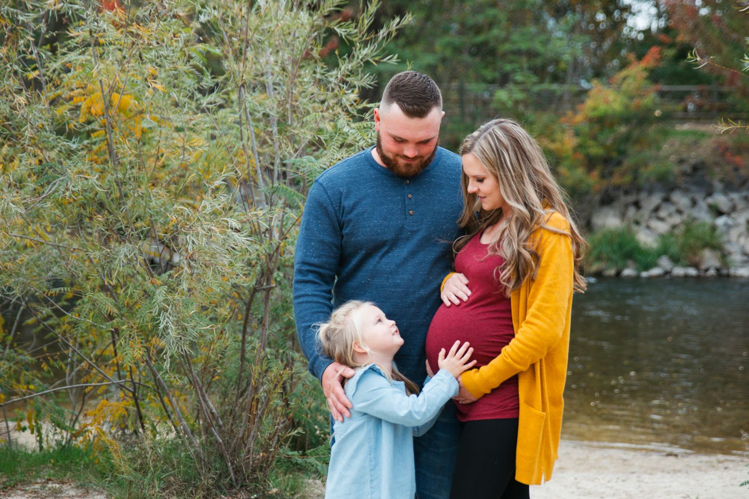 Family wearing coordinated autumn outfits poses together by a scenic river during fall foliage season - Maternity session in Eagle, Idaho