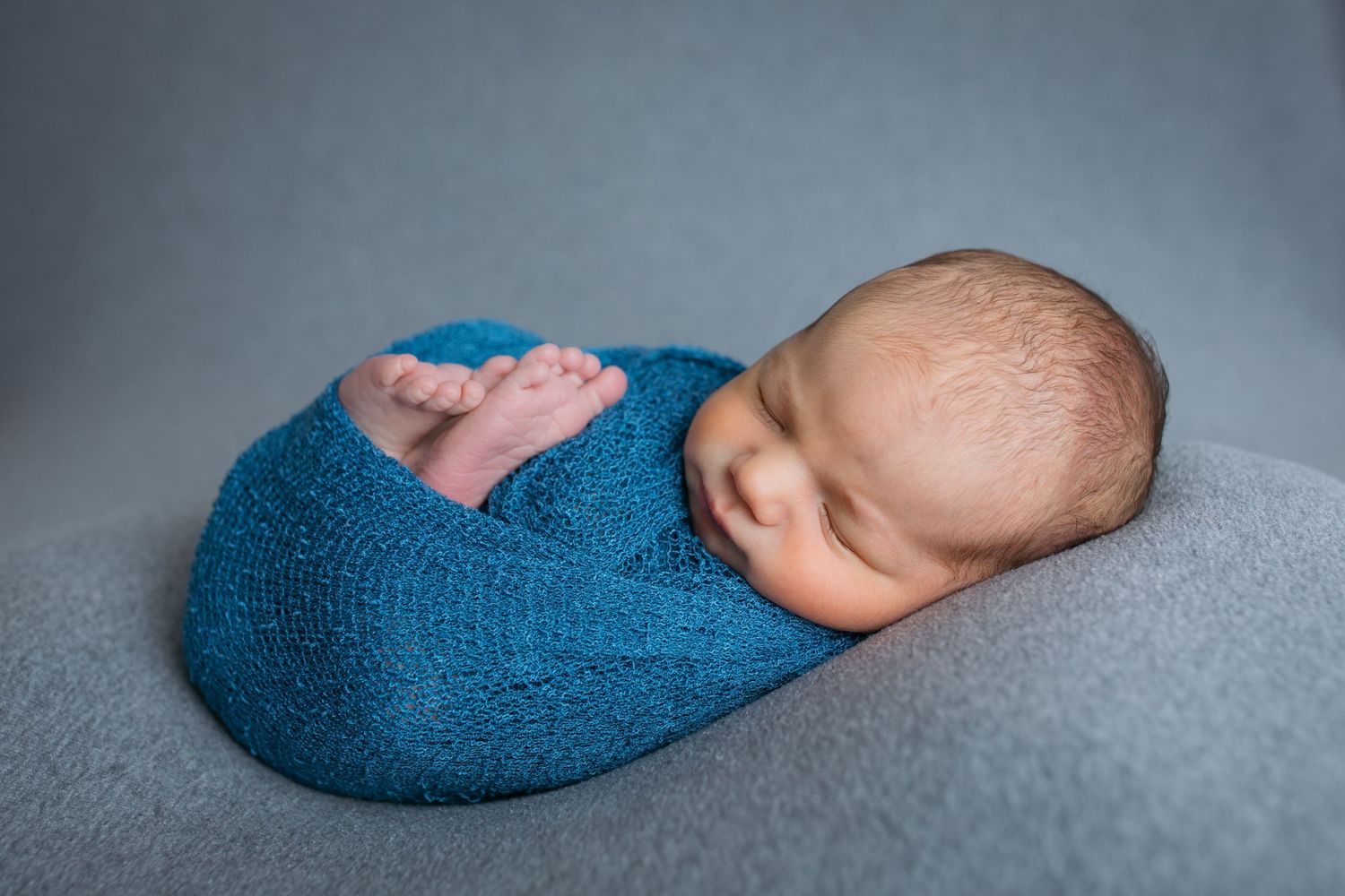 Newborn baby boy wrapped in blue swaddle sleeping peacefully on gray studio backdrop