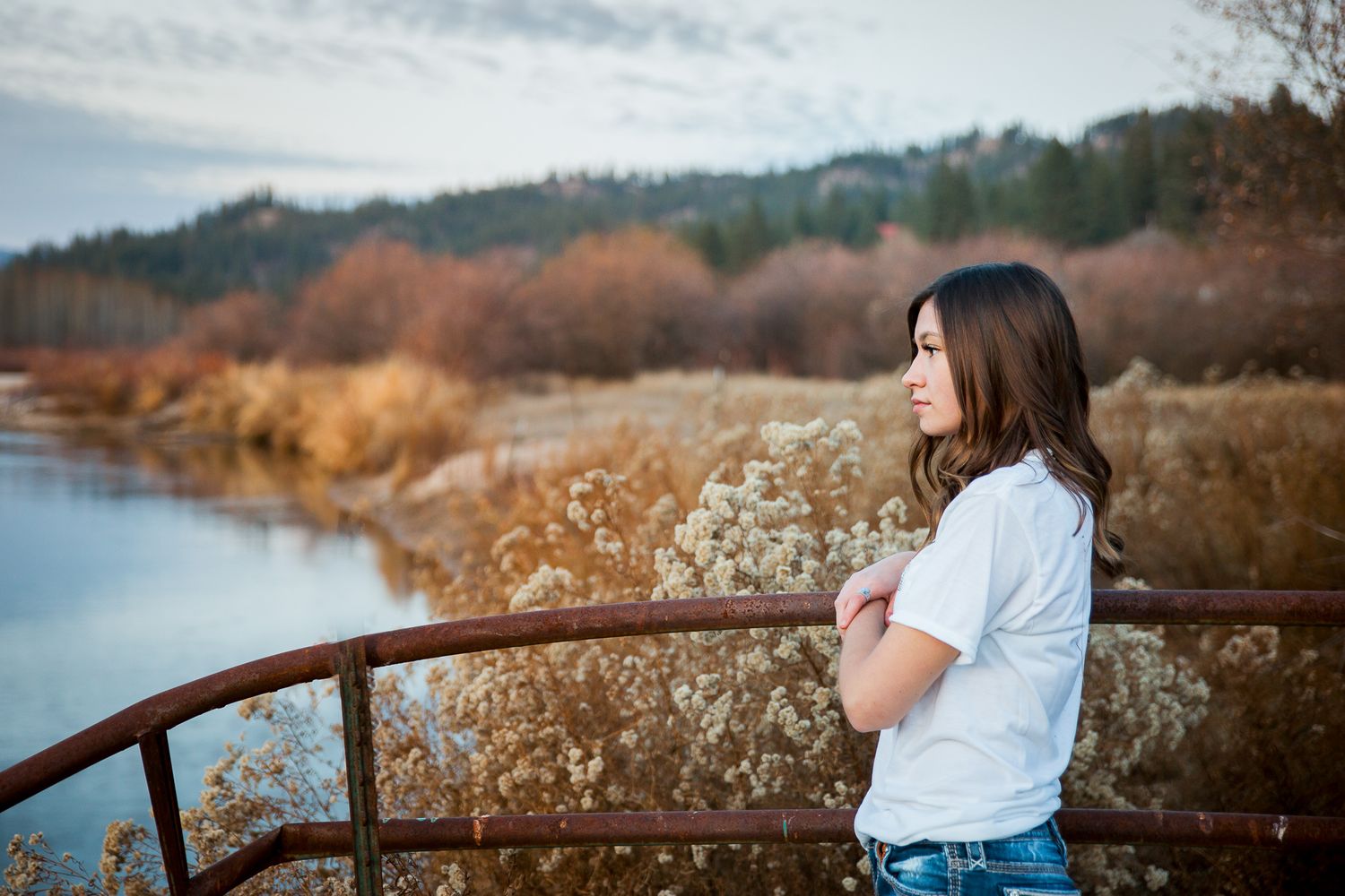 A person in a white shirt sits on a rustic railing near a lake during autumn with mountains in the background - Senior photos Boise Idaho