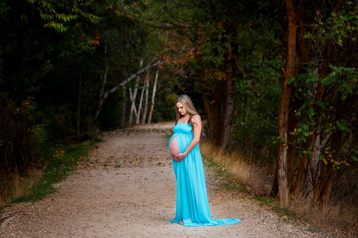 A woman in a flowing turquoise gown stands on a wooded path during a maternity photoshoot - Maternity session in Boise, Idaho