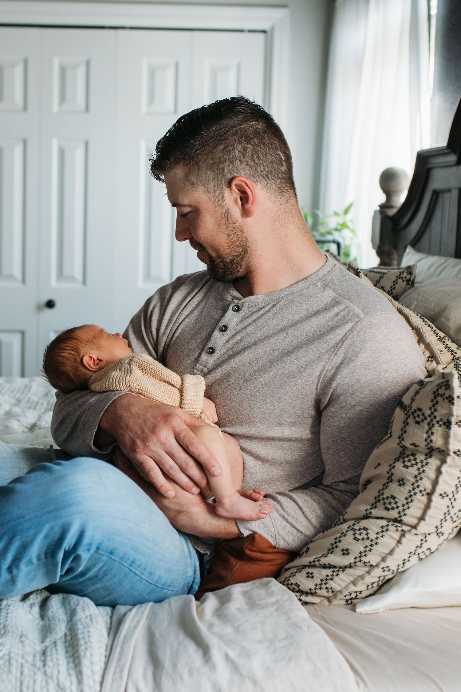 A dad relaxing together with a baby on a bed with patterned throw pillows in a bright, sunlit room - lifestyle in-home newborn photos Boise