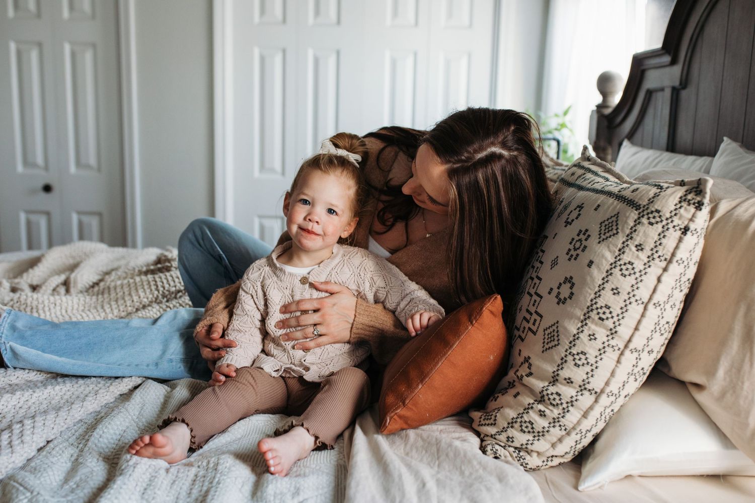 A baby lies on a bed wearing white clothes while being comforted by someone nearby - in-home lifestyle family photos Boise
