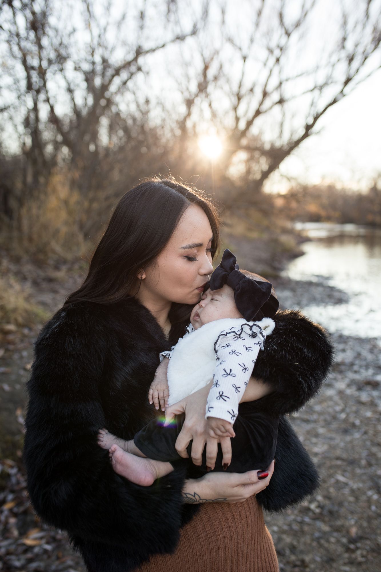 Mother holding and kissing her baby at Reid Merrill Park in Eagle, Idaho during a soft golden winter sunset by the river.