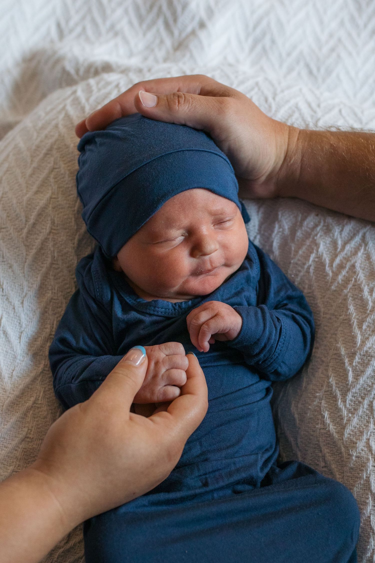 Gentle hands caress a sleeping newborn baby wearing cozy blue clothing on a textured white blanket. Fresh 48 session treasure valley idaho