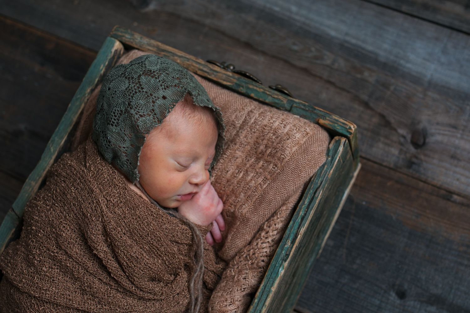 Sleeping newborn baby girl wrapped in earthy brown tones and posed in a rustic wooden box at a Nampa newborn photography studio.