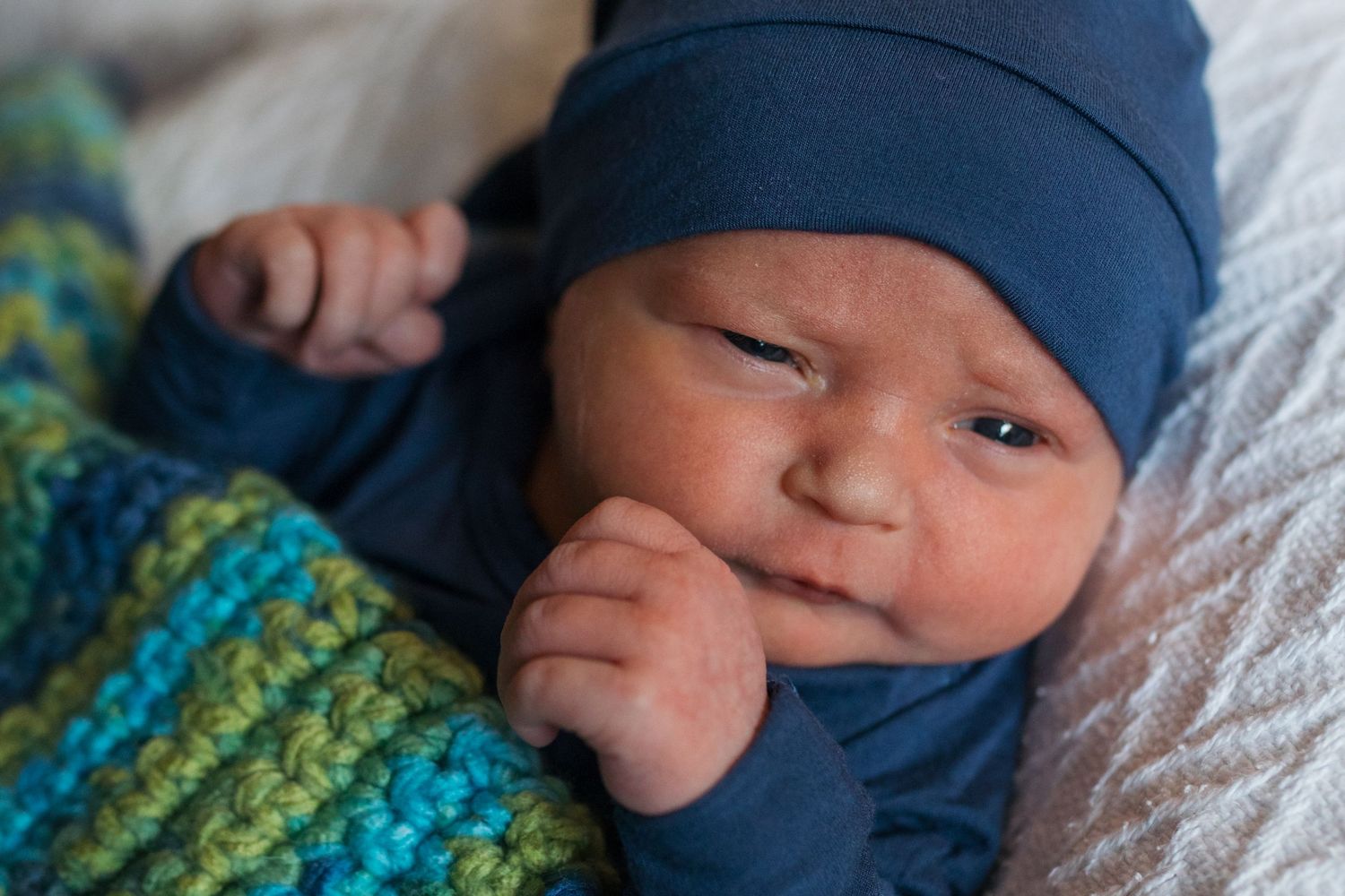 A baby wearing a navy blue hat lies on white bedding while wrapped in a green and blue striped blanket - Fresh 48 session in Boise, Idaho