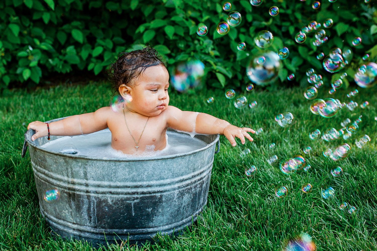 A baby splashes and plays in a metal tub outdoors surrounded by bubbles and green grass on a sunny day - 9 month milestone photos Boise