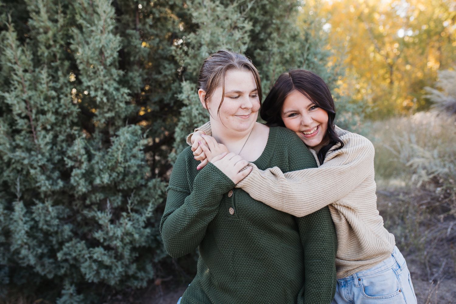 sisters hugging and laughing in outdoor setting with golden sunlight filtering through trees in the background. Boise foothills family photo