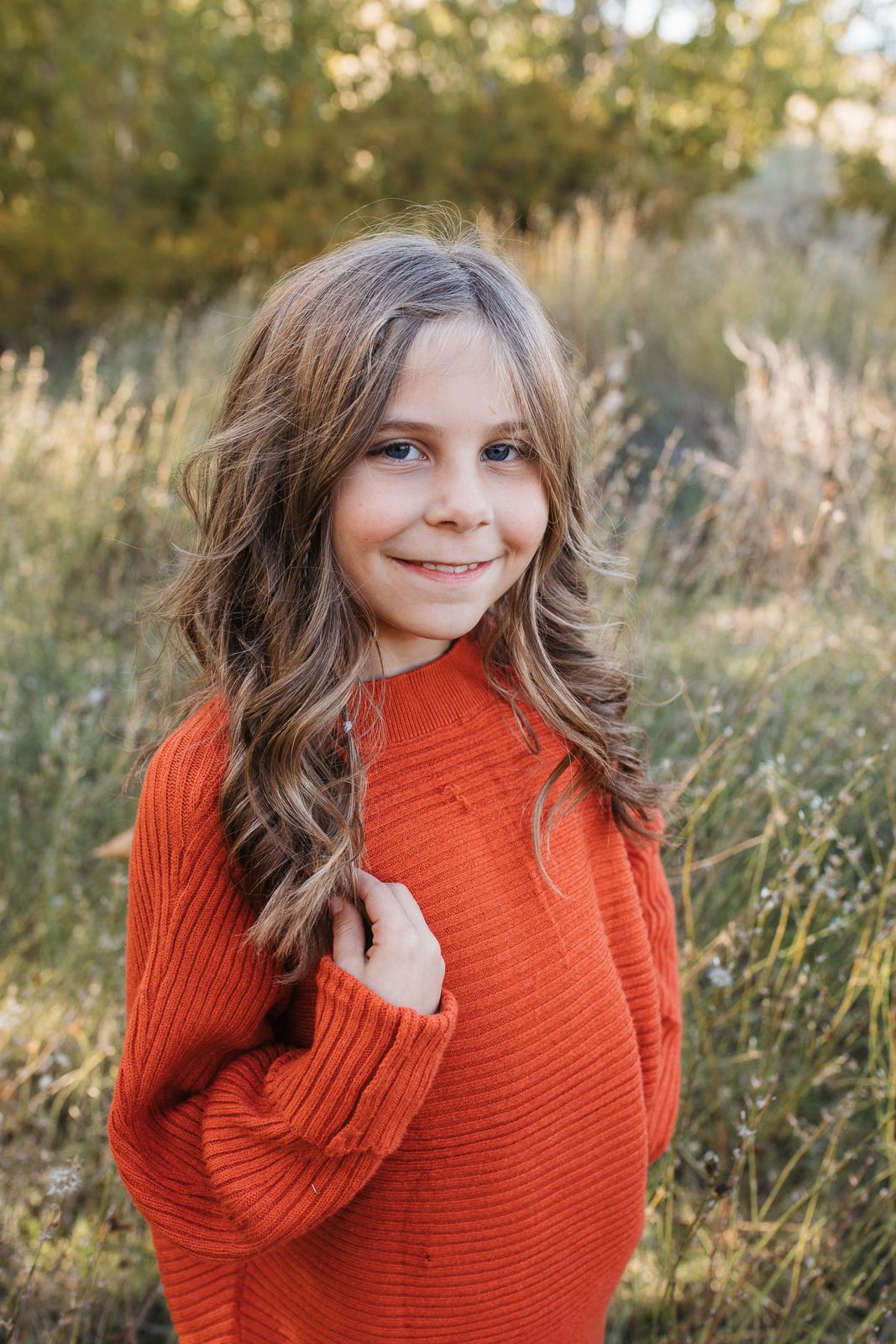A person wearing a cozy orange turtleneck sweater poses in a sunlit field of tall grass during golden hour. Boise foothills family photograp