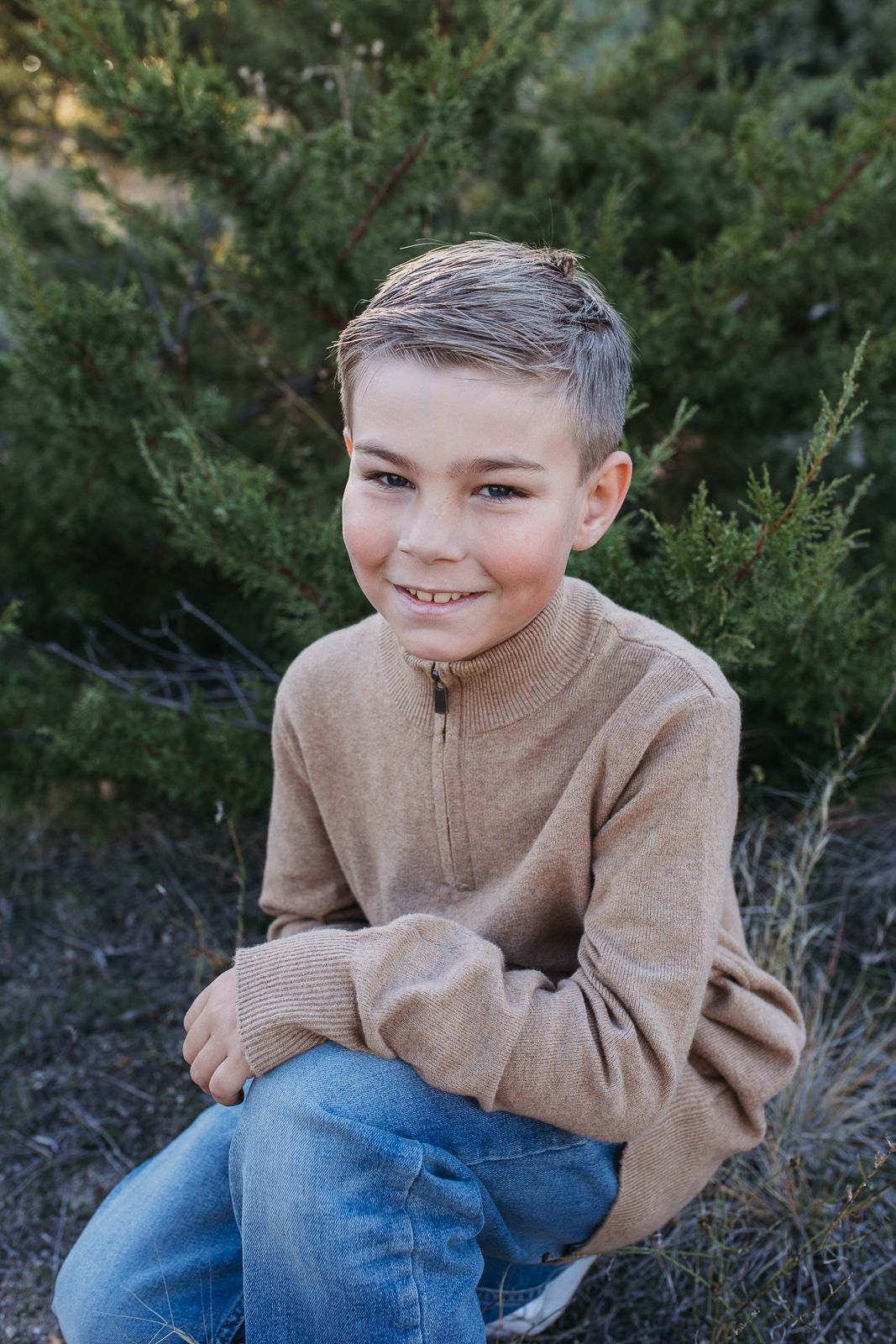 Smiling youth in tan sweater and jeans poses outdoors against green shrubs in natural portrait setting. Boise foothills family photography