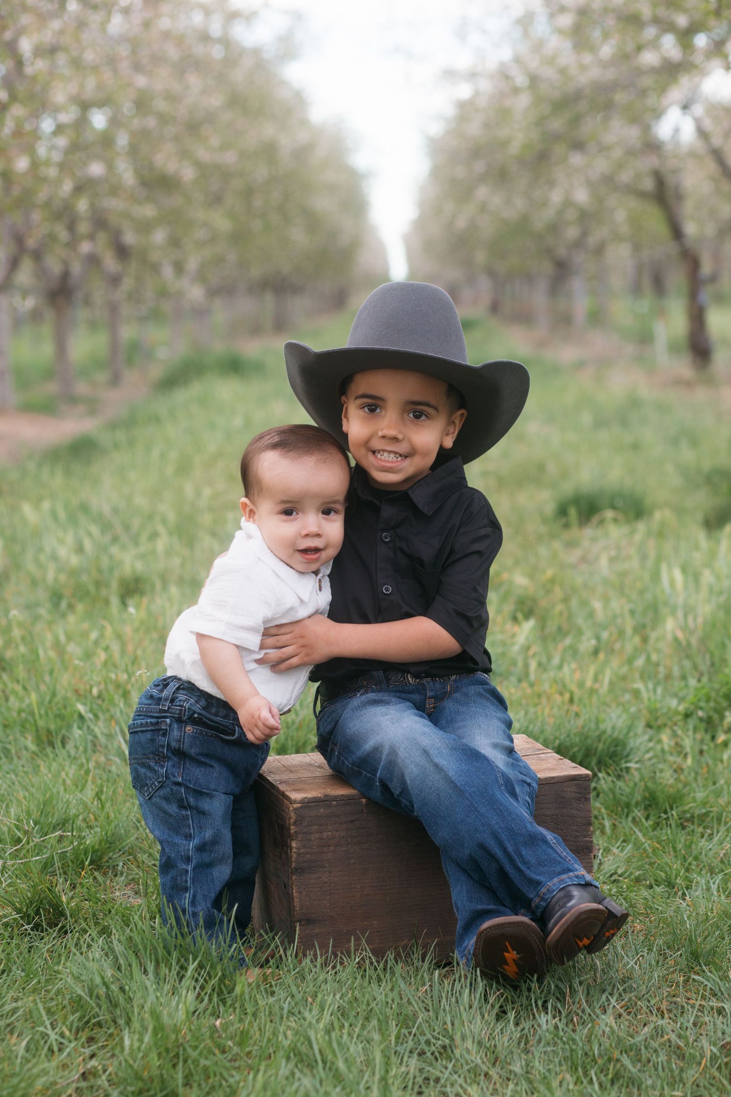 Young boy in cowboy hat holding baby sibling on bench in blossom orchard during spring session in Caldwell Idaho