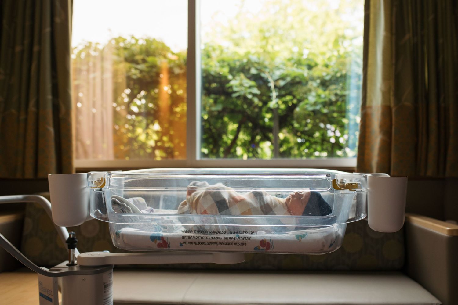 A newborn sleeps peacefully in a hospital bassinet by a window with natural light streaming in during the daytime.