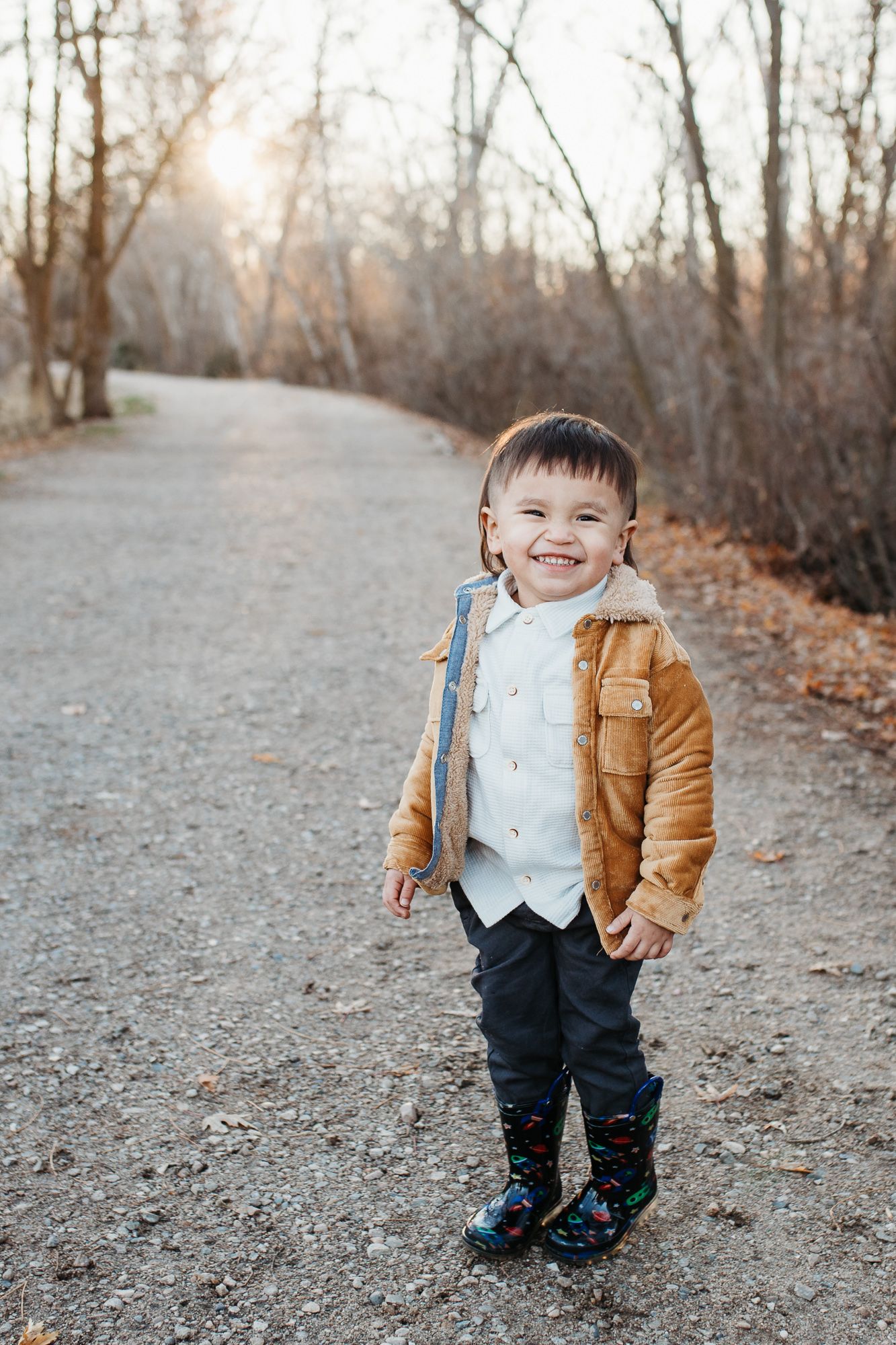 A child wearing a brown jacket and dark pants stands on a dirt path surrounded by bare autumn trees - lifestyle family photos in Boise