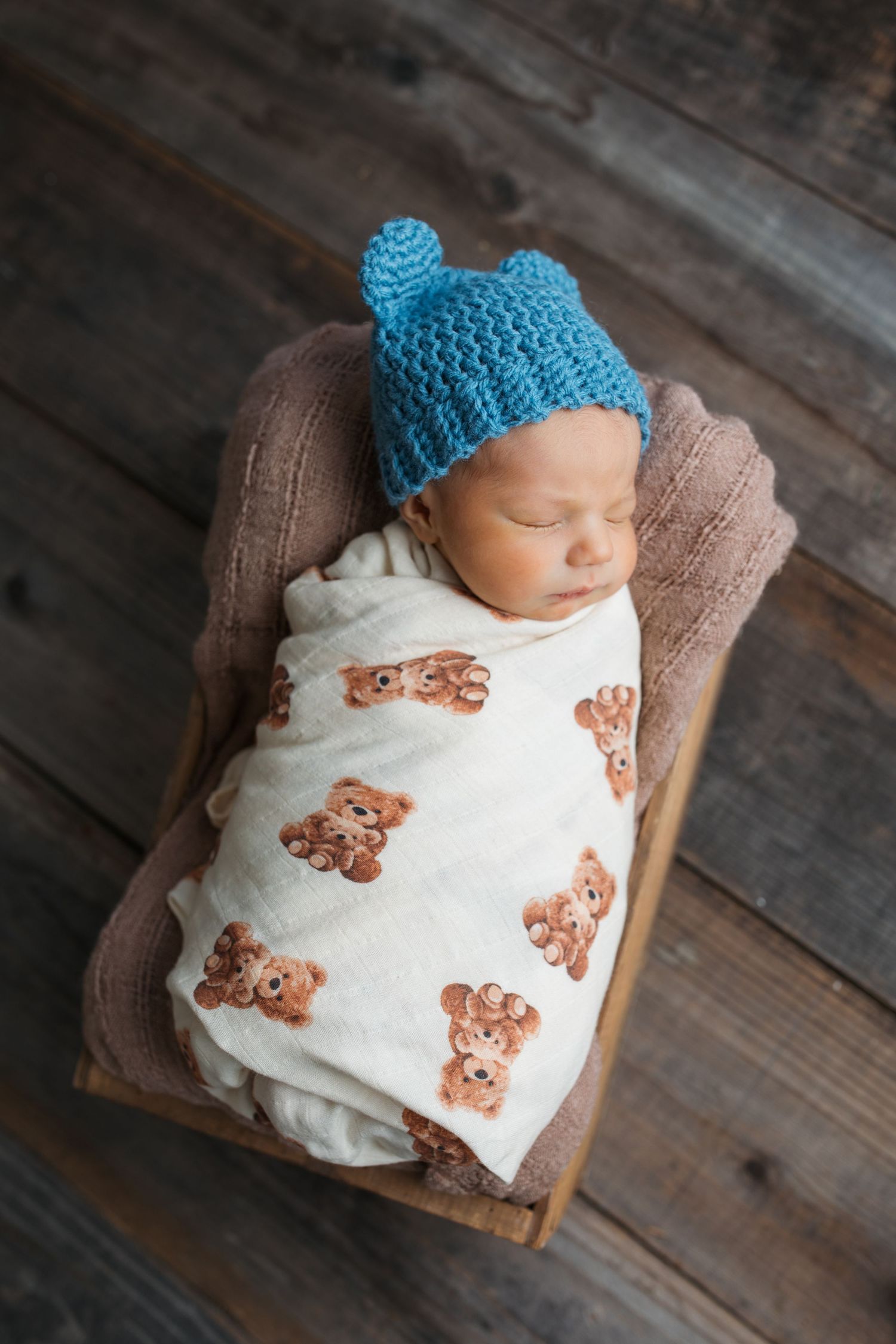 Newborn baby boy sleeping in wooden bed prop wearing blue knit hat and teddy bear swaddle