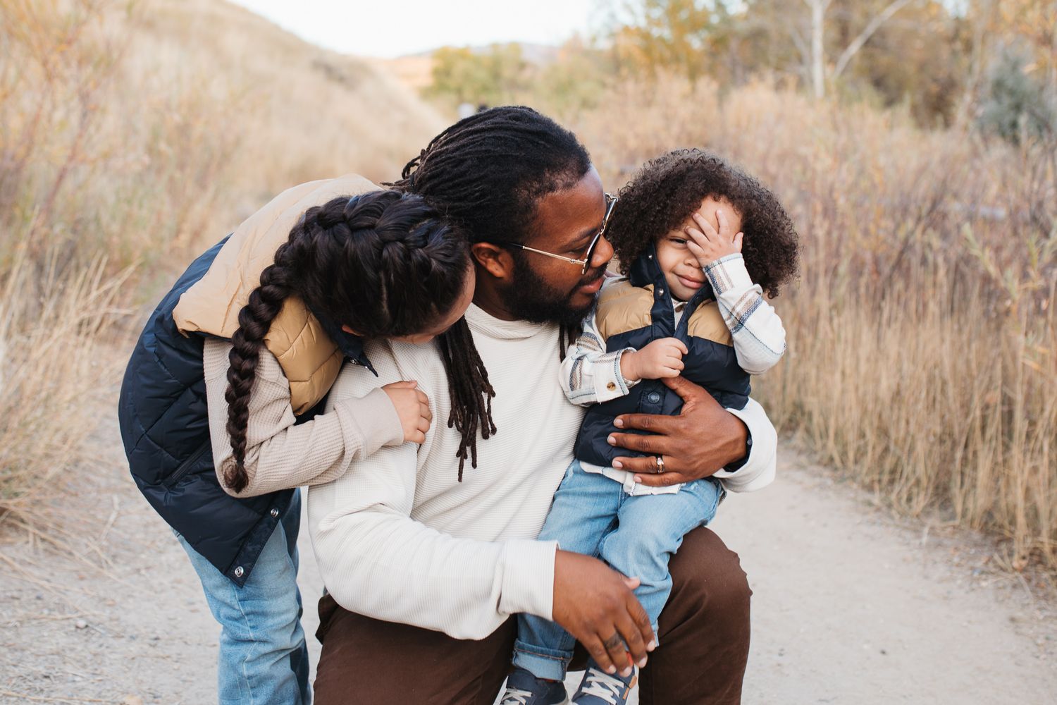 A family hug on a rustic dirt path surrounded by tall golden grass during sunset - lifestyle family photos in Boise