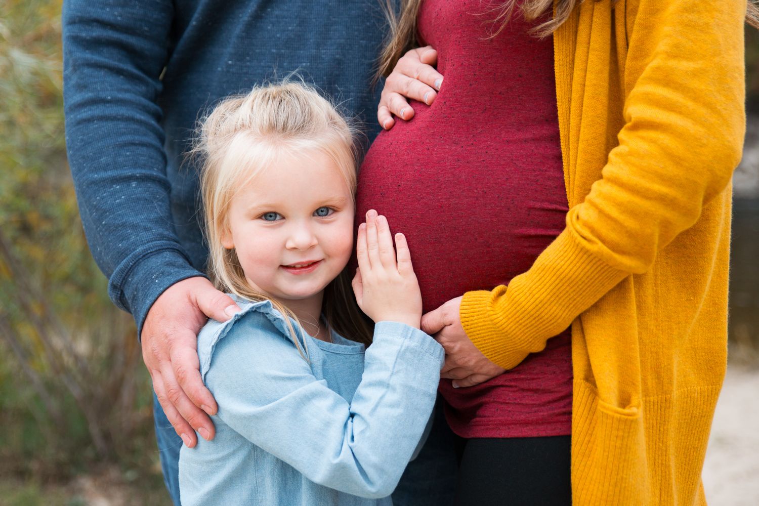 A pregnant woman in a burgundy dress stands with someone wearing a mustard yellow cardigan in an outdoor autumn setting - Maternity session