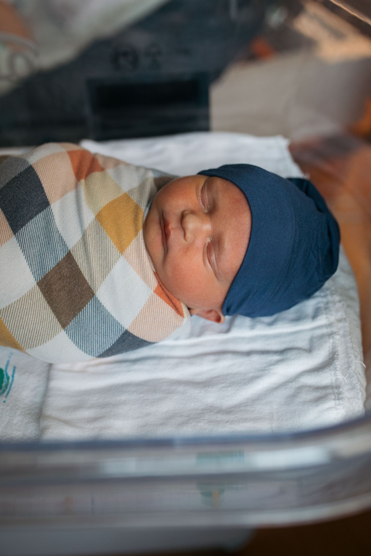 A sleeping newborn baby wearing a blue hat rests peacefully on white bedding with a geometric pattern blanket - Fresh 48 session in Boise