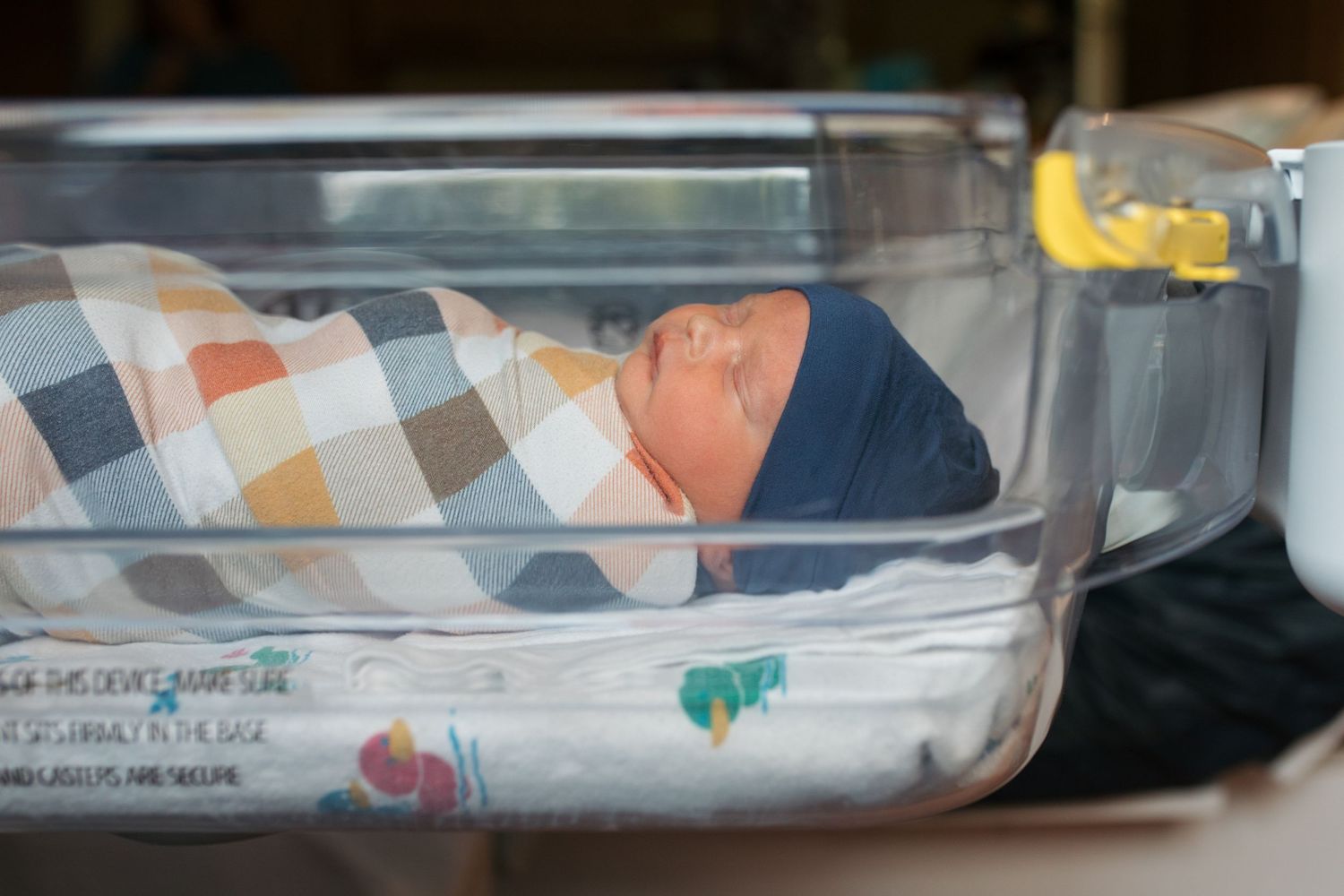 A newborn baby swaddled in a plaid blanket rests in a clear hospital bassinet. Fresh 48 session by Sara Elizabeth Photography