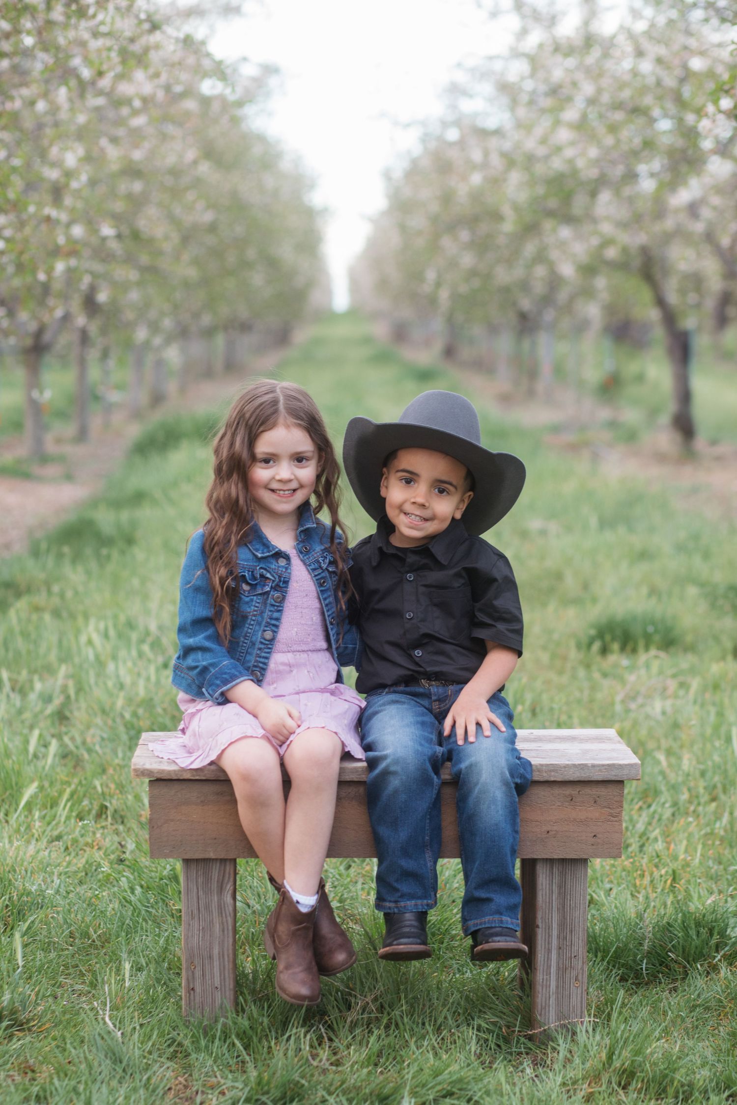 Brother and sister sitting on bench under spring blossoms during family session in Caldwell Idaho orchard