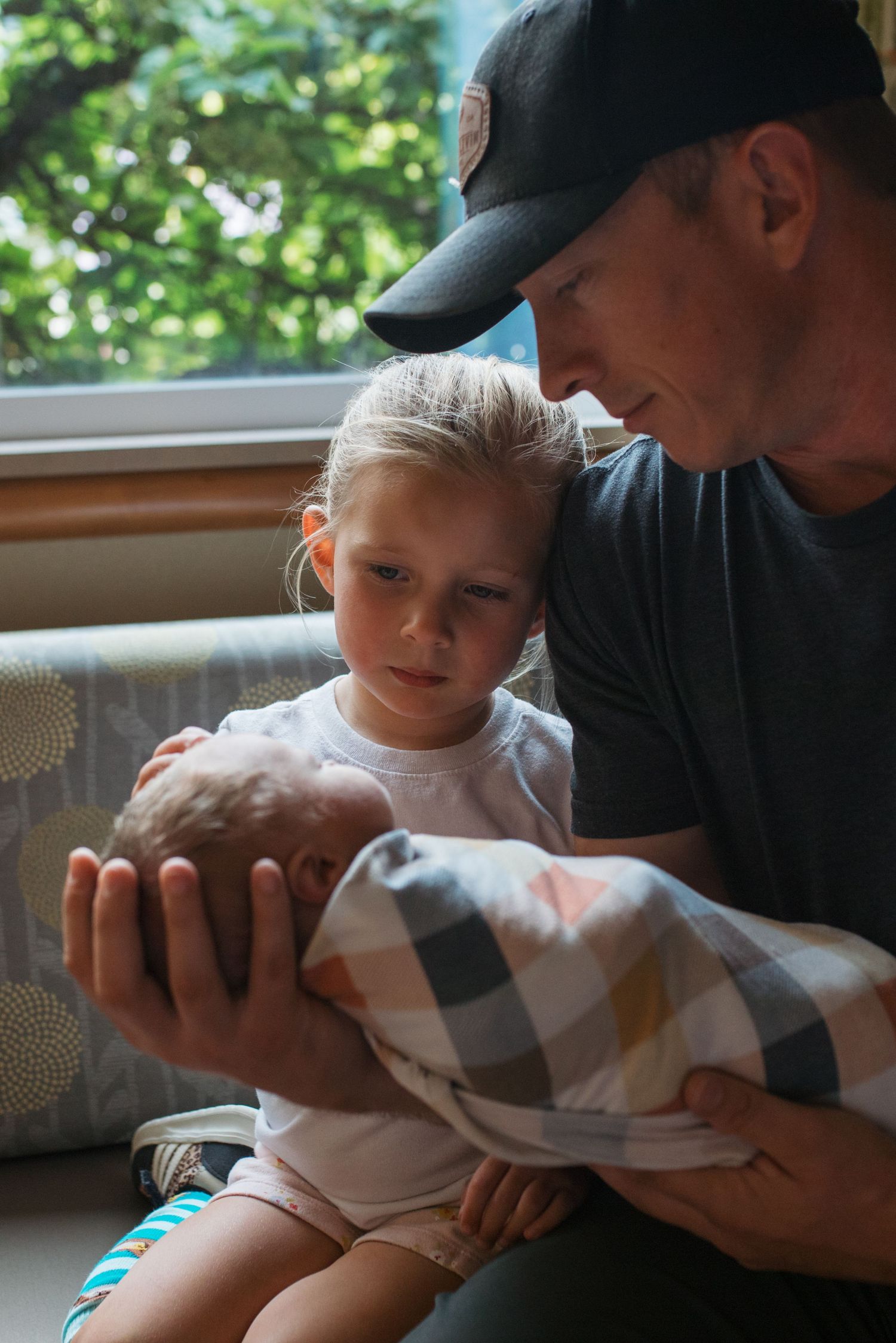 A tender moment by a window as a child and adult in a baseball cap look down at a newborn baby - Fresh 48 session in Boise, Idaho