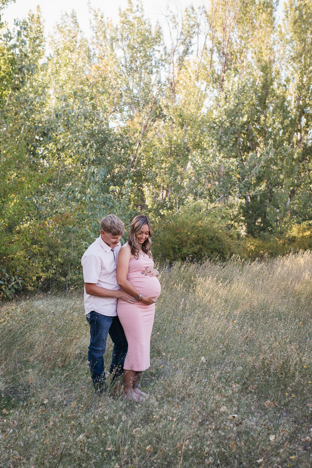 Pregnant woman standing in the foothills of Boise, holding her belly while her partner hugs her from behind - Maternity session in Boise