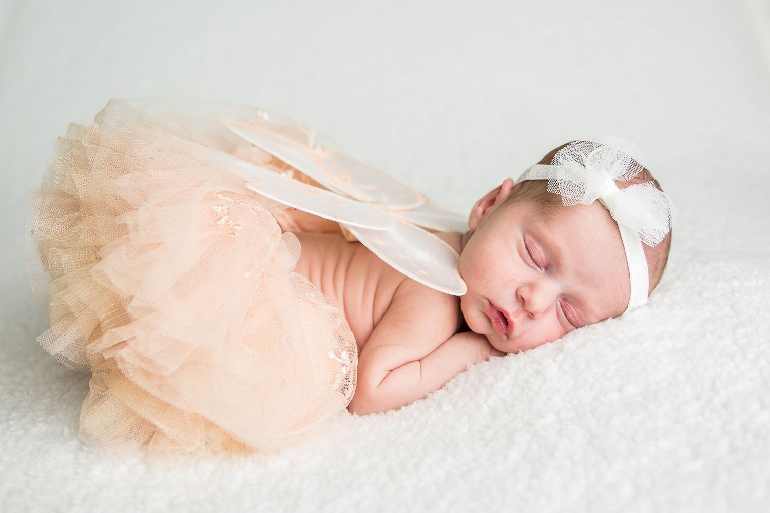 A sleeping newborn baby wearing a peach tulle tutu dress lies peacefully on white textured fabric studio newborn photography Boise Idaho
