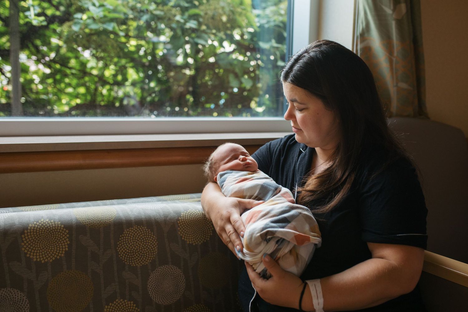 A person in a black shirt holds a small baby near a window with natural light streaming in - Fresh 48 session in Boise, Idaho