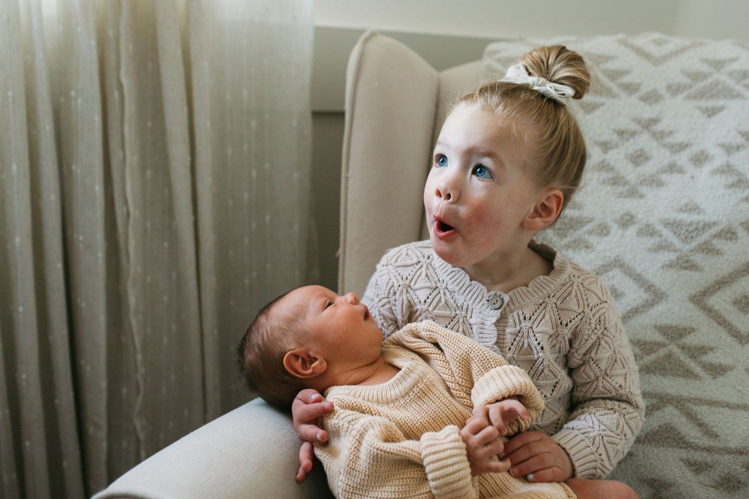 A toddler wearing a cream knit sweater sits and holds baby while looking upward with curiosity - lifestyle in-home newborn photos Boise