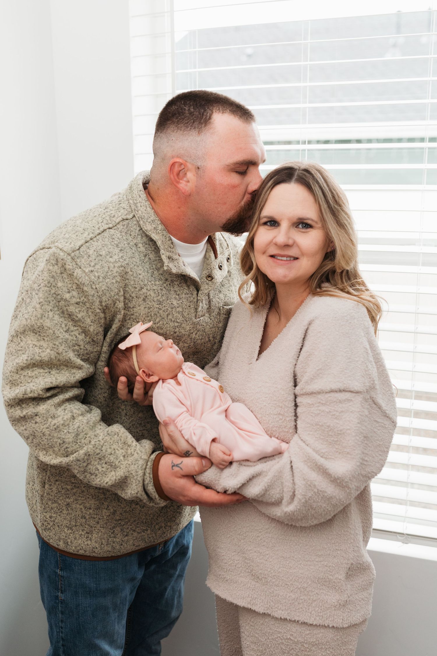 Parents holding their newborn daughter by a bright window, with dad kissing mom’s forehead during an in-home lifestyle newborn session idaho