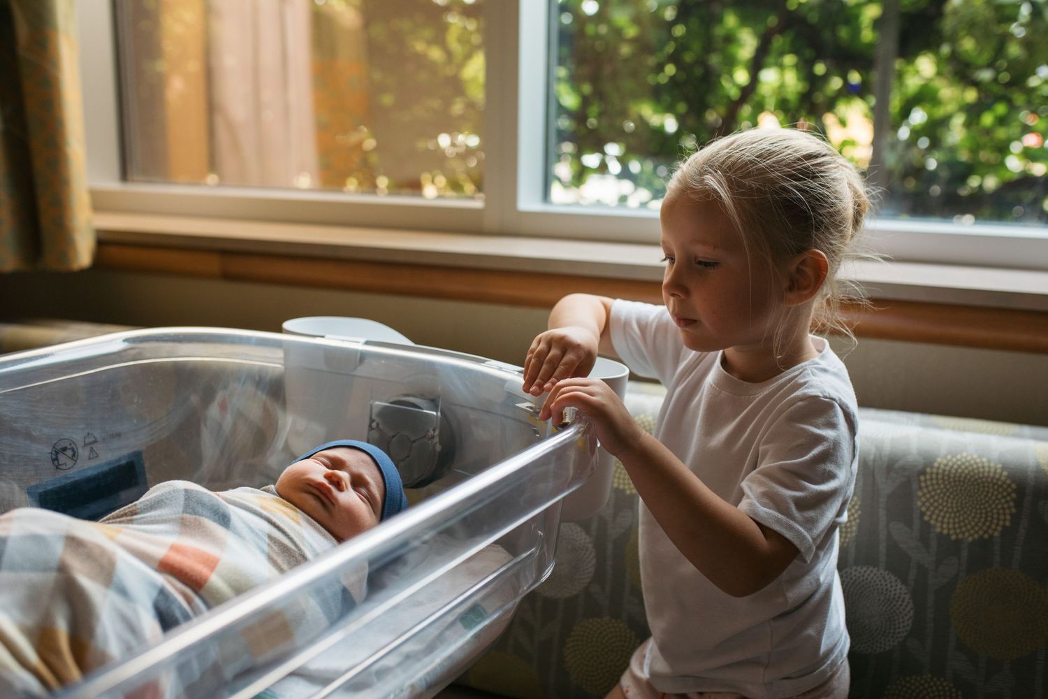 Fresh 48 newborn photo in Boise hospital of older sibling looking at baby in bassinet by the window