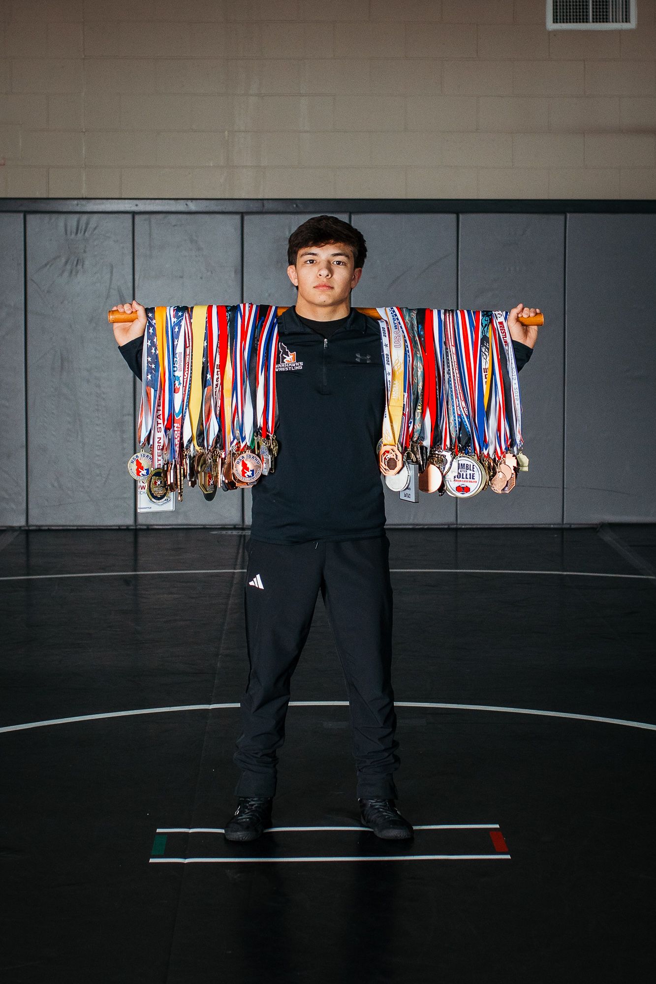 An athlete in black Adidas attire displays an impressive collection of colorful sports medals against a dark indoor backdrop - Senior photos