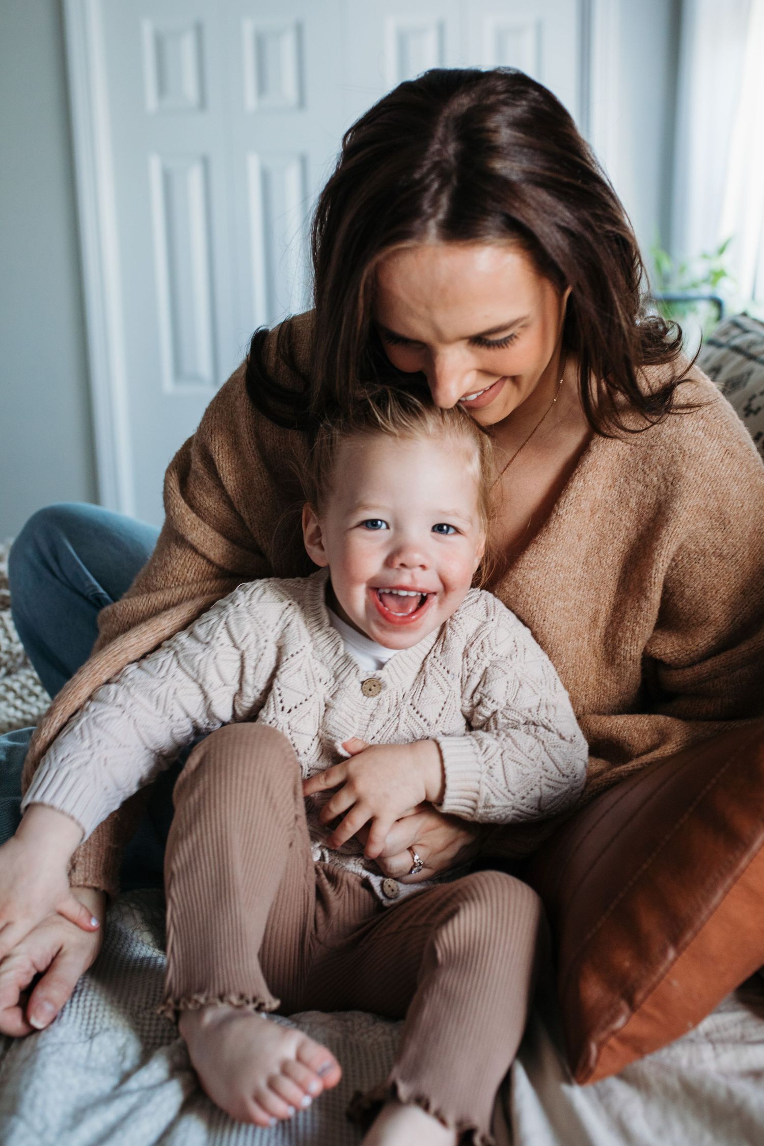 A parent and child share joyful moments together while relaxing on a bed in beige outfits - in-home lifestyle family photos Boise
