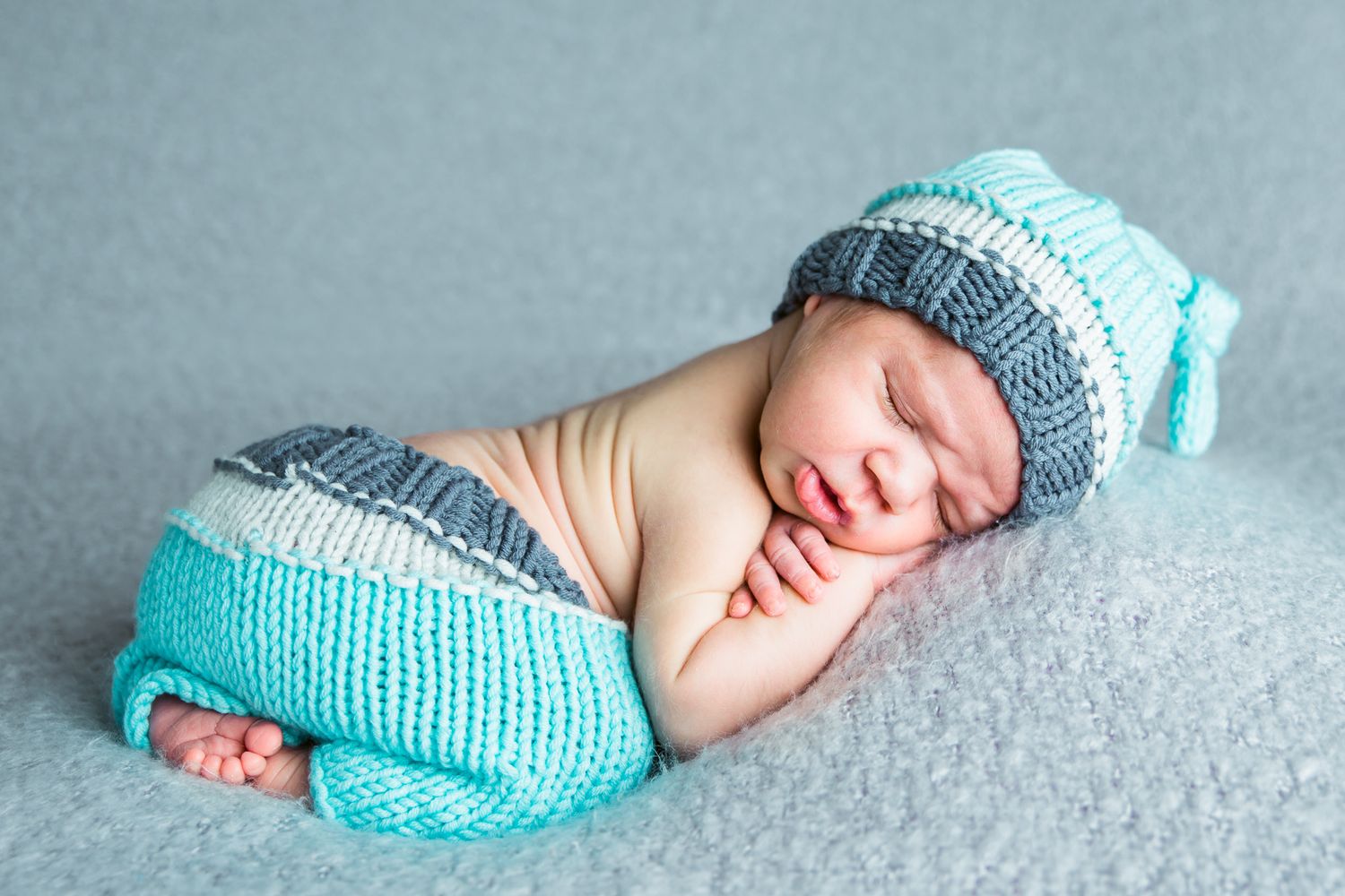 A sleeping newborn wearing a teal knitted hat and diaper cover lies on a soft gray blanket - studio newborn photography Nampa Idaho