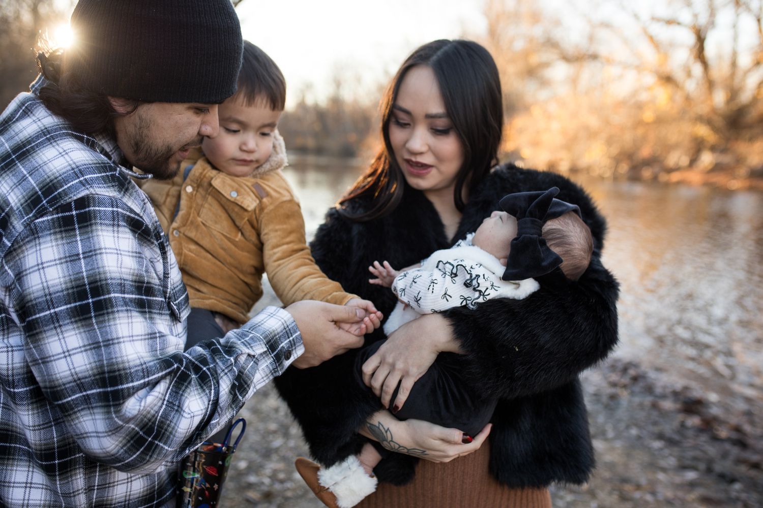 Parents holding their toddler and baby by the river at Reid Merrill Park in Eagle, Idaho during warm golden winter light.