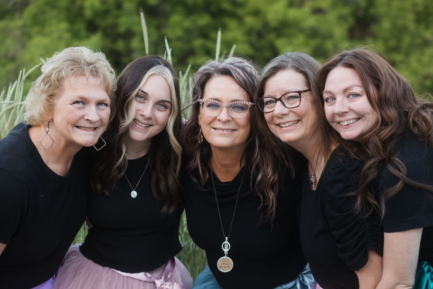Five sisters in black tops smiling together outdoors, posing for a group portrait with green foliage background.