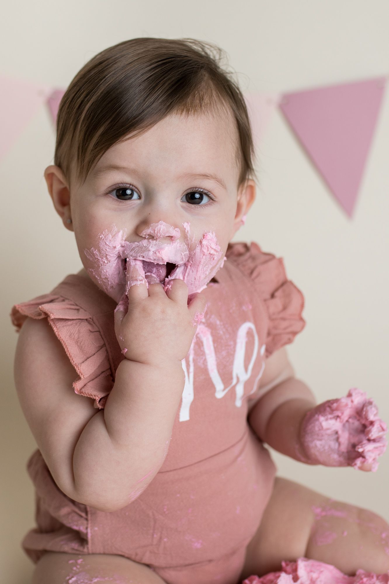 A baby in a pink shirt smiles while messily eating cake during their first birthday celebration - first birthday milestone photos Boise
