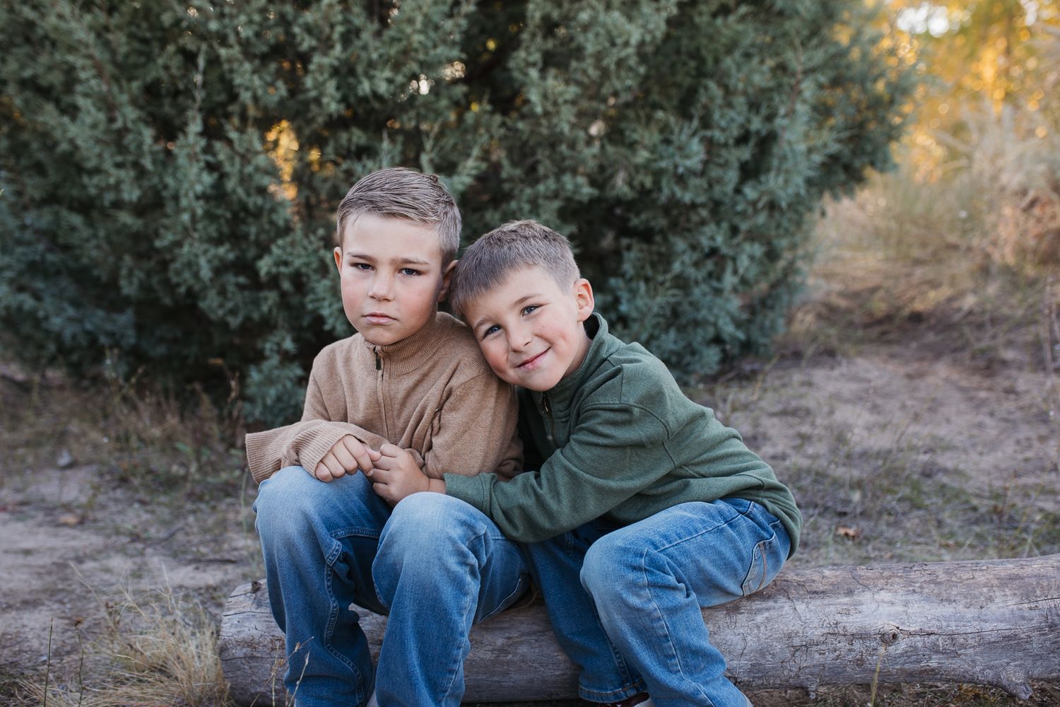 Two children in casual clothing sit close together on the ground near evergreen bushes on an autumn day. Boise foothills family photography