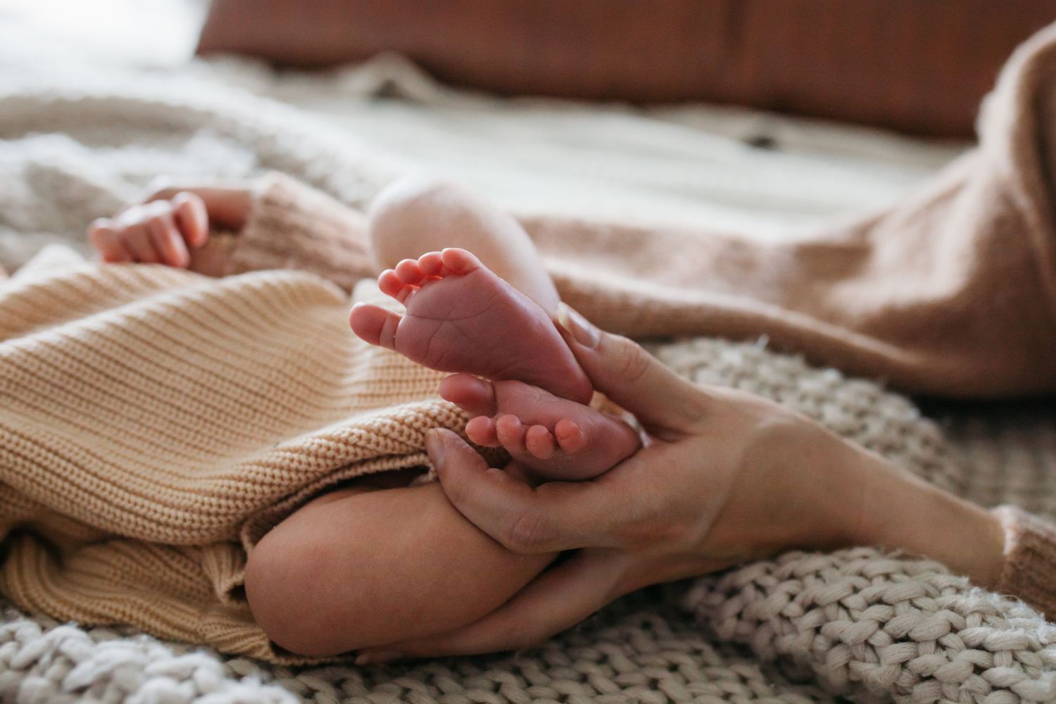 Gentle hands cradle a tiny newborn baby's feet against a soft knitted blanket in warm natural lighting - lifestyle in-home newborn photos