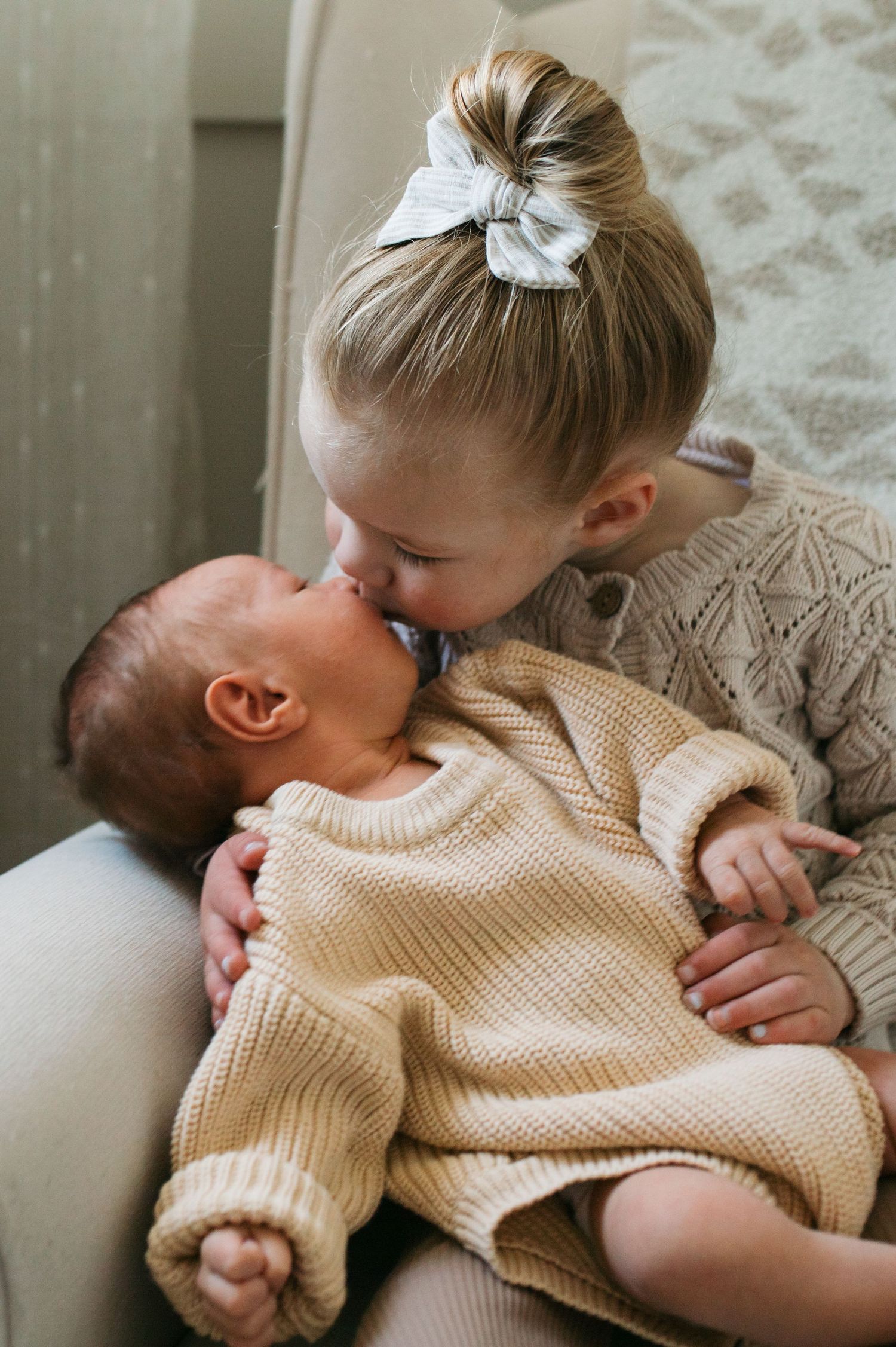 A tender moment between siblings as an older child in a knit sweater gives a gentle kiss to a baby wearing a beige sweater - newborn photos